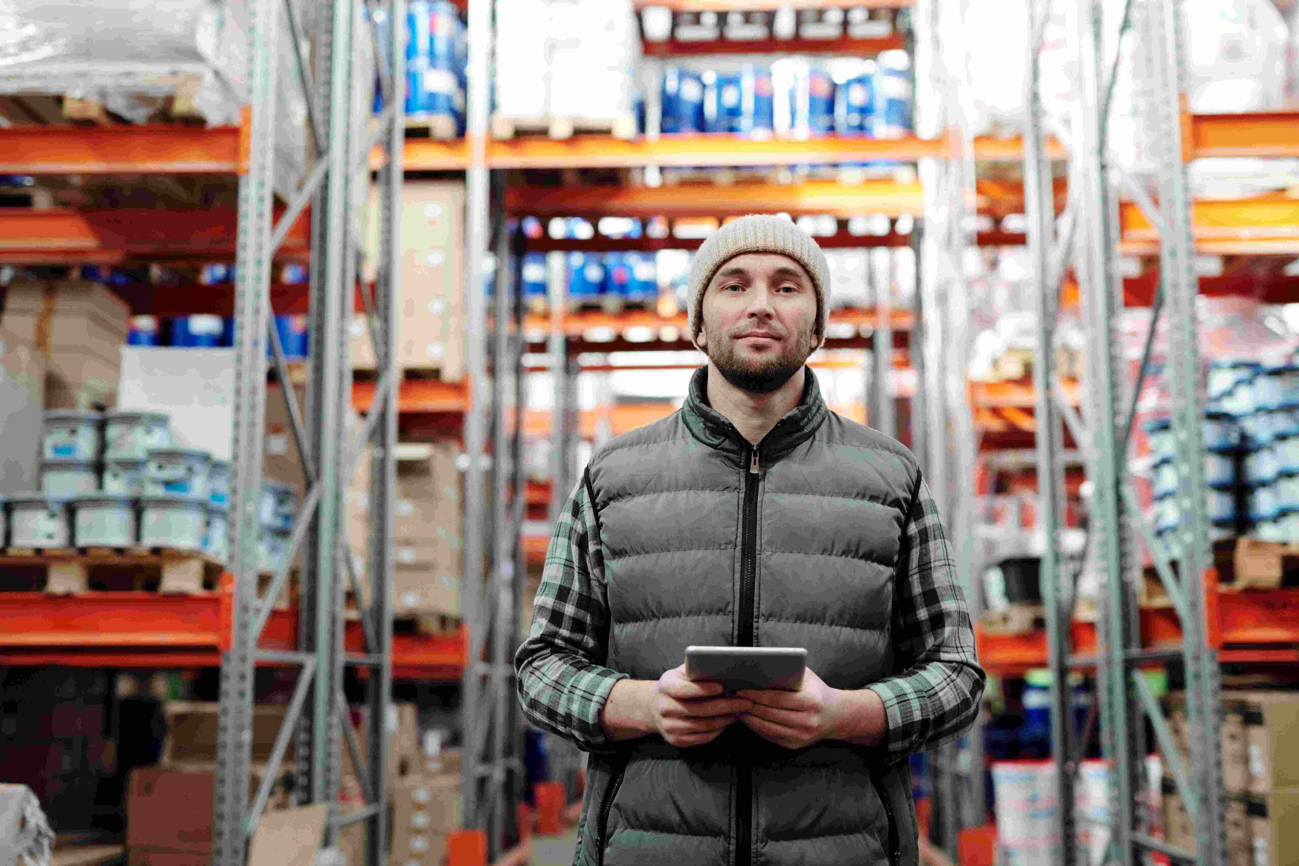 worker smiling in warehouse holding ipad with stretch wrapped pallets behind him